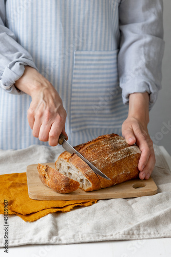 women\'s hands hold bread against the background of a light apron, a woman cuts bread, bread with a crispy crust is beautiful and appetizing, delicious bread lies on a wooden cutting board and a