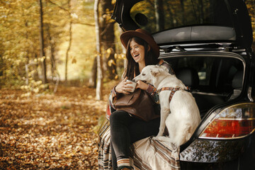 Sticker - Stylish woman traveller with backpack sitting with cute dog in car trunk in sunny autumn woods. Young hipster female traveling with swiss shepherd white dog. Travel and road trip with pet