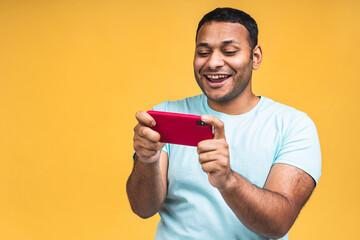 Portrait of a cheerful young african black indian man dressed in casual playing games on mobile phone isolated over yellow background.