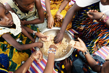 Group Of Black African Girls Sitting On A Mat, Dividing Their Frugal Meal, Eating With Their Hands From A Large Metal Plate