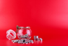 A Jar With Coins Inside And Many Coins Lying Outside The Jar Isolated On A Red Background.