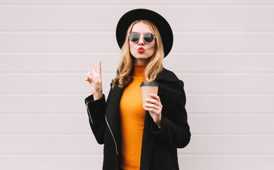 Canvas Print - Fashionable portrait of stylish young woman with coffee cup blowing her lips with red lipstick sending sweet air kiss showing peace hand sign wearing black coat and round hat on gray background