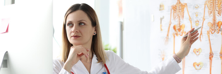 Medicine professor looking at computer screen and showing pen at poster with human skeleton