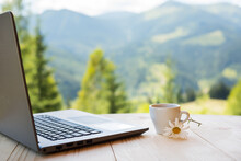 A Cup Of Coffee And A Laptop On A Wooden Table Outdoors Overlooking The Mountain Landscape.