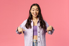 Portrait of curious good-looking, stylish female in denim jacket over crop-top, smiling excited and looking pointing fingers down at something interesting and unsual, stand pink background