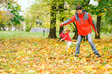 Man and dog wearing red similar coats playing together and having fun in autumn park