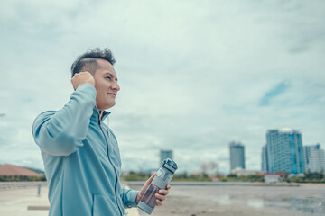 Wall Mural - The young athlete was listening to music and hold water bottle while running on the road near the beach and city background. Music and sport concept.