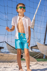 Handsome young boy in blue shorts and white shirt, wearing sunglasses standing on white sand on a volleyball beach court. High quality photo