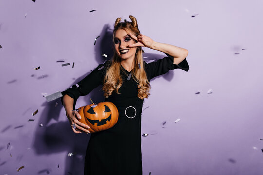 Cute caucasian woman in black dress posing after halloween masquerade. Indoor photo of smiling cheerful girl with pumpkin.