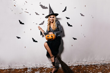 Wall Mural - Positive long-haired woman in halloween costume laughing to camera. Refined blonde girl in witch hat posing with bats on background.