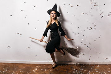 Wall Mural - Full-length portrait of appealing blonde woman posing in studio in halloween. Attractive witch in black dress sitting on magic broom.