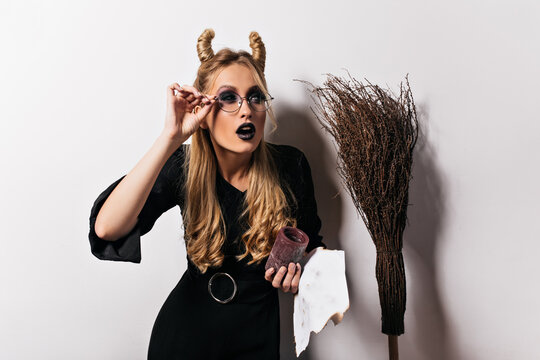 Attractive female wizard fooling around during halloween photoshoot. Blonde caucasian lady in witch costume posing on white background.