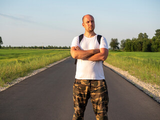 A middle-aged man is standing on an asphalt road with a backpack on his back. Hands folded on his chest. Travel concept