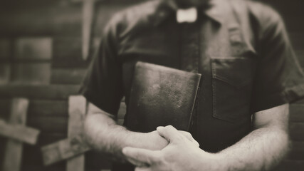 Clergyman, reverend or priest wearing a clerical collar and clutching a bible. Preacher preaching the gospel in front of an old rustic rural church or cemetery.