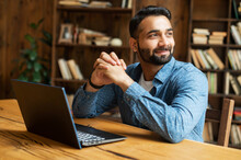Happy And Optimistic Indian Man In Casual Wear Using Laptop In Modern Office, Successful Inspired Arabic Male Entrepreneur Looks Away And Smiles Thoughtfully, Planning Next Step