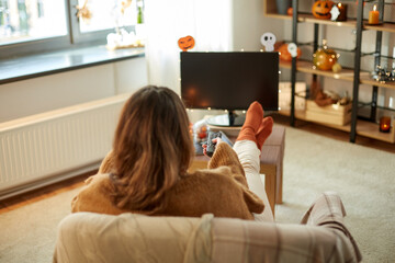 Poster - halloween, holidays and leisure concept - young womanwith remote control watching tv and resting her feet on table at cozy home