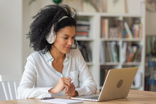 Portrait Of African American Woman Talking On Video Conference Call Using Laptop And Headphones Taking Notes On Notepad. Brunette Sits At Table In Home Office. Close Up.