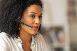 © kinomaster - Portrait of a young African American woman talking on video conference call using headset. Brunette with curly hair in white blouse sits at table in light home office. Close up.