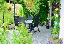 Beautiful Outdoor House Patio With Stone Carpet Flooring, Growing Green Flowers And Plants, And Table With Chairs. House Terrace Overgrown With Plants With Garden Furniture.