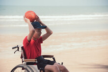 Happy Disabled Teenage Boy On Wheelchair Playing A Ball, Activity Outdoors With Father On The Beach Background, People Having Fun And Diverse People Concept.