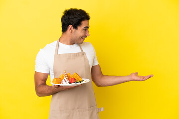 Sticker - Restaurant waiter holding waffles over isolated yellow background with surprise expression while looking side