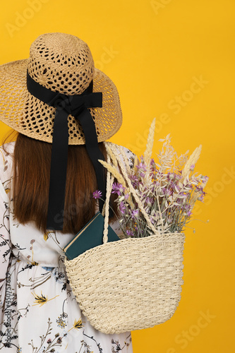 Woman holding beach bag with beautiful bouquet of wildflowers and book on yellow background, back view