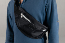Closeup Studio Shot Of Male Model In Blue Long Sleeve Shirt Hanging Trendy Urban Small Black Crossbody Strap Casual Fanny Pack Bag In Front Gray Background