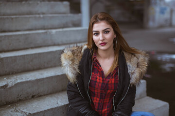 Poster - Young Caucasian lady sitting on stairs wearing makeup, a winter coat, and a shirt