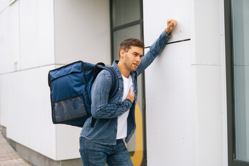 Wall Mural - Medium shot of overworked delivery service employee having painful infarction stopping by wall and holding heart in city street. Exhausted food delivery man with large thermo backpack outdoors.