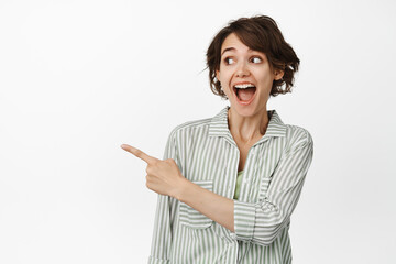 Wall Mural - Portrait of excited caucasian woman laughing, smiling amazed and pointing finger left, showing advertisement, standing over white background