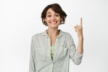 Wall Mural - Portrait of excited smiling woman, pointing finger up and looking happy, demonstrating advertisement, click on link, standing against white background