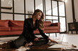 © Look! - Curly attractive woman in stylish black silk home suit sits on carpet in living room, holds phone and works in laptop.