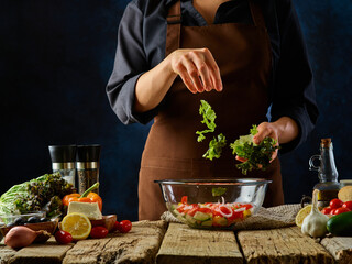 The chef tosses pieces of greens into the Greek salad. Lots of colorful ingredients on a wooden table. Delicious vegetarian dish. Diet food, few calories. Restaurant, cafe, hotel, home cooking.