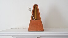 An Old Fashioned Metronome Beating At A Largo Pace On A White Shelf.