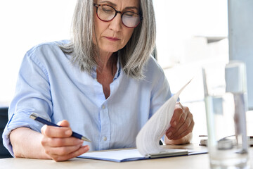 Focused concentrated older businesswoman in glasses reading signing legal document partnership contract sit at table in office. Executive ceo signing bank financial loan investment agreement.