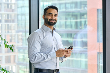 Smiling indian ceo businessman holding using cell phone mobile apps standing in office near panoramic window looking at camera. Digital tech apps and solutions for business corporate development.