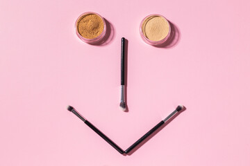 Wall Mural - Funny face with make-up brushes and mineral powder on pink background with copy space, top view. Cosmetics humor and beauty concept.