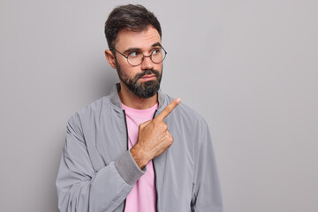 Wall Mural - Serious self confident bearded man shows advertisement copy space demonstrates promo deal points at upper right corner wears round spectacles jacket isolated over grey background. Look at this