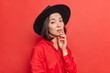 © WHstudio Leushin N - Elegant charming young Asian lady keeps hand on chin looks with serious confident expression has natural dark hair healthy skin wears black hat red shirt poses against bright background indoor