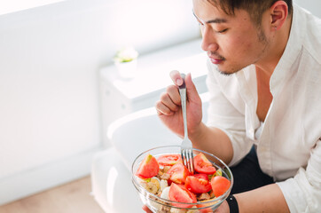 Portrait of young handsome Chinese man eating a fresh, delicious and healthy tomate salad with tune on the sofa in the living room of a minimalist and clean home