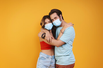 Poster - Beautiful young couple in protective face masks embracing while standing against yellow background