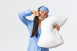 Smiling pleased cute asian girl in blue pyjama and sleeping mask, hugging pillow and stretching hands delighted as finally going bed, want sleep or waking up in morning, white background