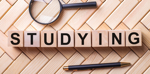 Wooden cubes with the word STUDYING stand on a wooden background between a magnifying glass and a pen