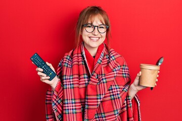 Canvas Print - Redhead young woman wrapped in a red warm red blanket eating icecream and watching tv smiling with a happy and cool smile on face. showing teeth.