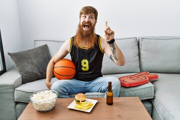 Wall Mural - Caucasian man with long beard holding basketball ball cheering tv game smiling amazed and surprised and pointing up with fingers and raised arms.