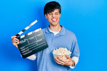 Poster - Handsome hipster young man eating popcorn holding cinema clapboard sticking tongue out happy with funny expression.