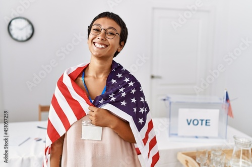 Young hispanic woman with short hair at political campaign election holding usa flag looking positive and happy standing and smiling with a confident smile showing teeth