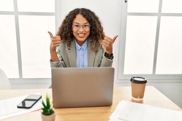 Wall Mural - Beautiful hispanic business woman sitting on desk at office working with laptop success sign doing positive gesture with hand, thumbs up smiling and happy. cheerful expression and winner gesture.