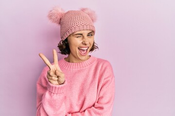 Wall Mural - Young brunette woman wearing cute wool cap smiling with happy face winking at the camera doing victory sign. number two.