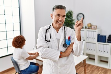 Canvas Print - Middle age doctor man at the clinic with a patient with a big smile on face, pointing with hand and finger to the side looking at the camera.